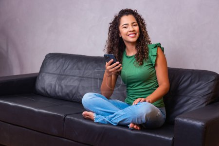 Téléchargez les photos : Jolie jeune femme africaine reposer sur le canapé utiliser téléphone intelligent sourire regarder le chat à l'écran avec petit ami lire agréable sms message nouvelles médias en ligne, technologie moderne tous les jours utilisation pour le plaisir ou concept d'affaires - en image libre de droit
