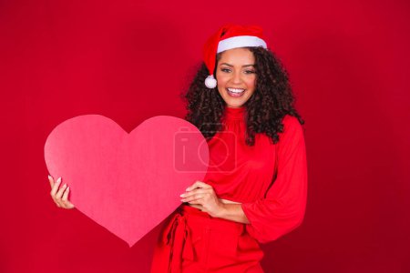 Foto de Sonriente joven afro chica en el sombrero de Navidad sosteniendo una tarjeta de corazón enorme con espacio libre para el texto. - Imagen libre de derechos