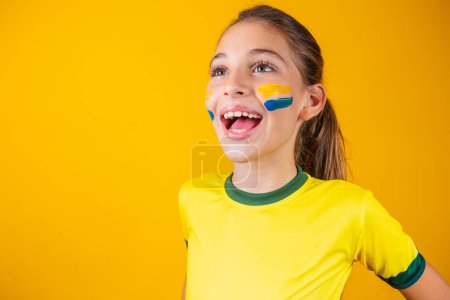 Téléchargez les photos : Supporteur de football, équipe du Brésil. Belle petite fille acclamant pour son équipe sur fond jaune - en image libre de droit