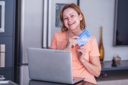 Téléchargez les photos : Femme âgée tenant une carte de travail et un ordinateur brésiliens. Femme âgée tenant un cahier d'exercices avec concept de bureau à domicile. - en image libre de droit