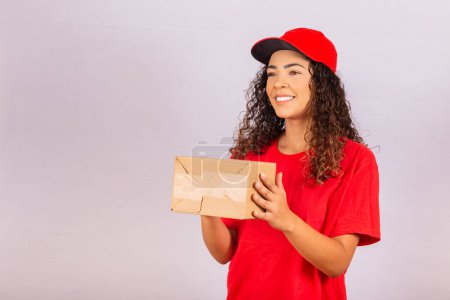 Photo for Beautiful young messenger delivering a package. A delivery woman in a red uniform is smiling with a package in her hands. Online shopping. Equality of women. - Royalty Free Image