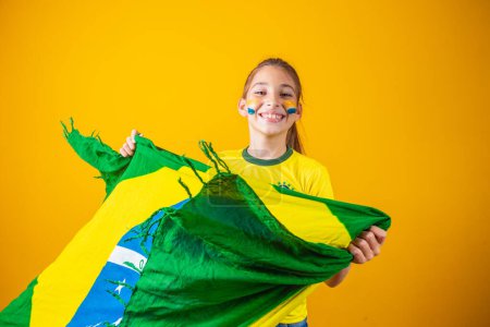 Téléchargez les photos : Petite fille caucasienne avec drapeau brésilien sur fond jaune. Enfant supporter brésilien - en image libre de droit