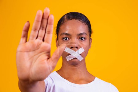 Téléchargez les photos : Femme noire sur fond jaune avec la bouche fermée en silence. Notion de préjudice, d'abus et de racisme - en image libre de droit