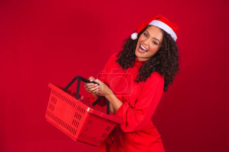 Téléchargez les photos : Afro femme souriant tenant panier vide regardant la caméra souriant portant santa claus chapeau. Shopping de Noël - en image libre de droit