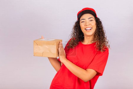 Photo for Beautiful young messenger delivering a package. A delivery woman in a red uniform is smiling with a package in her hands. Online shopping. Equality of women. - Royalty Free Image