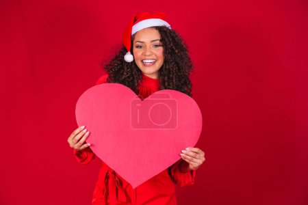 Foto de Sonriente joven afro chica en el sombrero de Navidad sosteniendo una tarjeta de corazón enorme con espacio libre para el texto. - Imagen libre de derechos