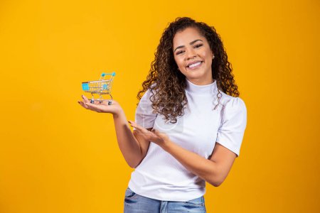Photo for Girl with small shopping card. Smiling and dancing curly hair woman in shopping concept. Young woman with a miniature cart. E-commerce and business. Shopping car. Woman shopper. Yellow background. - Royalty Free Image