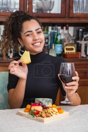 Foto de Mujer joven bebiendo vino y comiendo queso sola en la sala de estar. Mujer comiendo bocadillos de queso y bebiendo vino para relajarse después del trabajo. - Imagen libre de derechos