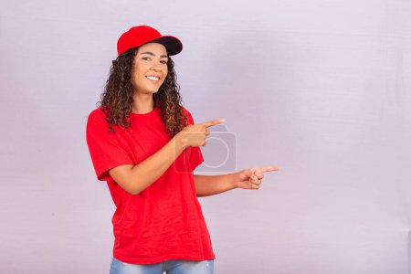 Photo for Delivery employee woman in red cap blank t-shirt uniform work courier in service during quarantine coronavirus covid-19 virus, pointing fingers aside on workspace isolated on white background studio - Royalty Free Image
