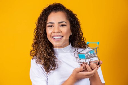 Photo for Smiling curly hair woman in shopping concept. A young woman with a miniature cart. E-commerce and business. Commercial vehicle. Woman buyer. Yellow background. - Royalty Free Image
