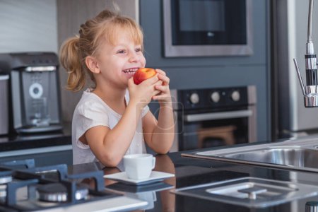 Photo for Little blonde girl eating an apple in the kitchen - Royalty Free Image