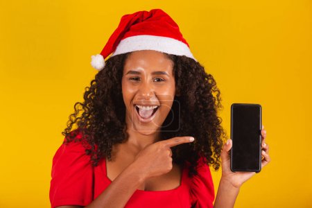 Téléchargez les photos : Souriant jeune Santa femme en chapeau de Noël tenant téléphone portable avec écran vide vide isolé sur fond jaune. - en image libre de droit