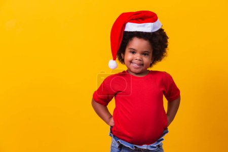Téléchargez les photos : Petite fille afro en t-shirt rouge et chapeau de Père Noël habillé pour Noël. Campagne de Noël et Nouvel An - en image libre de droit