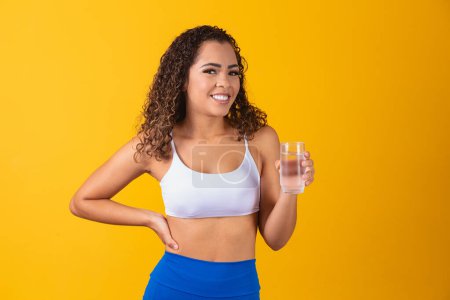 Foto de Mujer afro joven bebiendo agua en fondo amarillo. Chica joven con vaso de agua. Una joven bebiendo agua del vaso. Concepto de hidratación - Imagen libre de derechos
