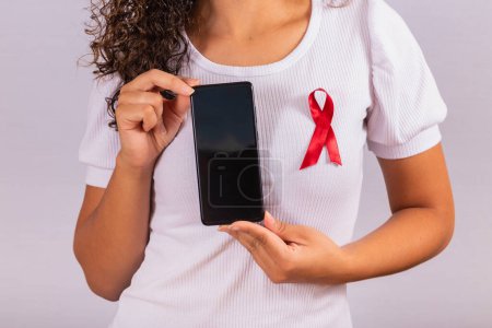 Foto de El concepto del Día Mundial del SIDA. Mujer joven sostiene teléfono inteligente con pantalla en blanco y cinta roja en la camiseta con el concepto de prevención y programación del VIH. - Imagen libre de derechos