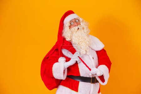 Téléchargez les photos : Père Noël avec un énorme sac dans la hâte de livrer des cadeaux de Noël. Père Noël sur fond jaune - en image libre de droit