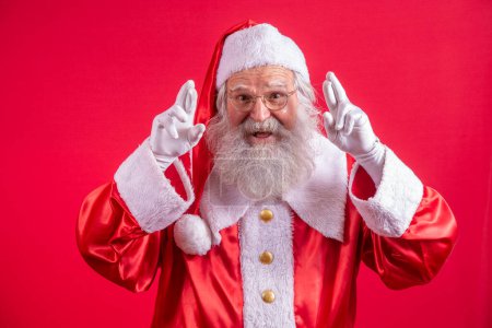 Photo for Portrait of dreamy elderly man in santa claus costume  crossing fingers making a wish, christmas magic, winter holidays. Indoor studio shot isolated on red background - Royalty Free Image