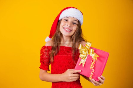 Photo for Cute cheerful girl in a Christmas hat on a colored background holding a gift - Royalty Free Image