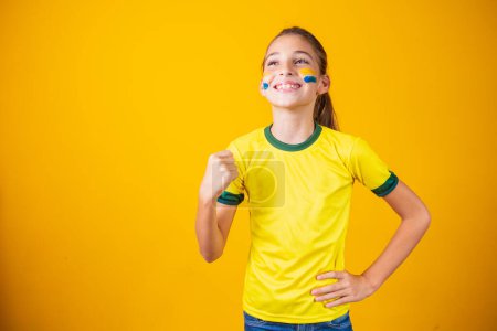 Téléchargez les photos : Belle petite fille enracinant pour son équipe sur fond jaune. Petite fille célébrant le but et la victoire du Brésil - en image libre de droit