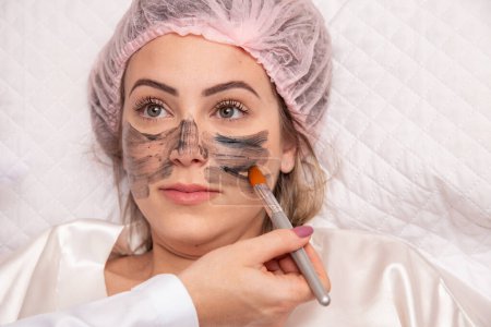 Photo for Woman is lying in the cosmetologist s office on the procedure of moisturizing her face. The cosmetologist applies a moisturizing mask to the patient's face. - Royalty Free Image