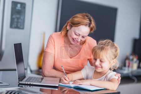 Photo for Smiling mature mother helps her daughter in preparing chores at home. Online education concept. - Royalty Free Image