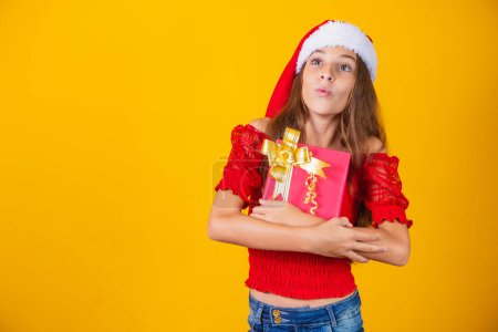 Téléchargez les photos : Mignonne jolie fille gaie en chapeau de Noël sur fond coloré tenant un cadeau - en image libre de droit
