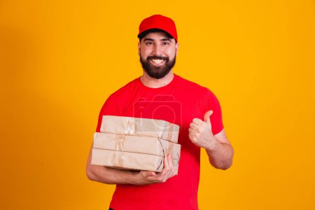 Photo for Best Service Concept. Smiling caucasian delivery man in red cap and stacking holding stack of cardboard boxes while standing isolated over yellow studio background. Mail carrying packages - Royalty Free Image