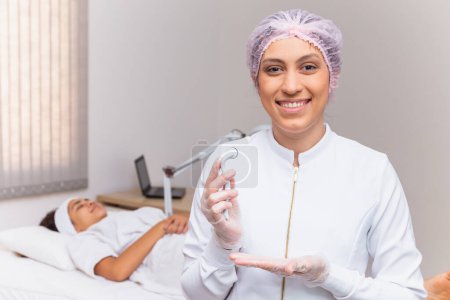 Photo for Closeup on beautician holding microneedling smiling at a camera with the patient in the background - Royalty Free Image