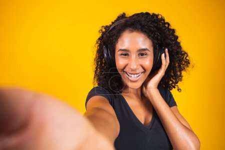Téléchargez les photos : Jeune femme africaine portant des écouteurs faisant un selfie sur fond jaune. - en image libre de droit