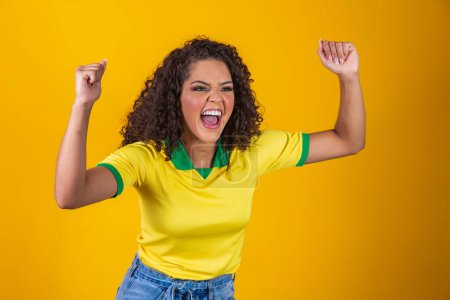 Téléchargez les photos : Soutien du Brésil. Brésilienne fan de cheveux bouclés célébrant sur le football, match de football sur fond jaune. Couleurs Brésil. - en image libre de droit