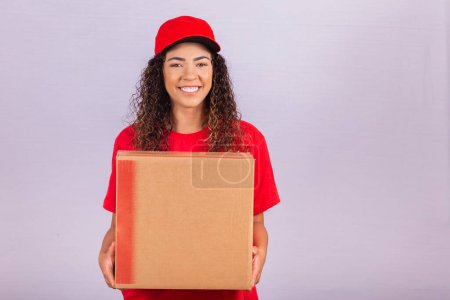 Photo for Beautiful young messenger delivering a huge package. A delivery woman in a red uniform is smiling with a large cardboard box in her hands. Online shopping. Equality of women. - Royalty Free Image