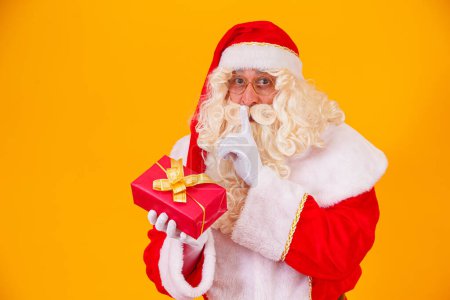 Téléchargez les photos : Père Noël sur fond jaune tenant un cadeau dans les mains et faisant un signe de silence avec l'autre main. La surprise du Père Noël - en image libre de droit