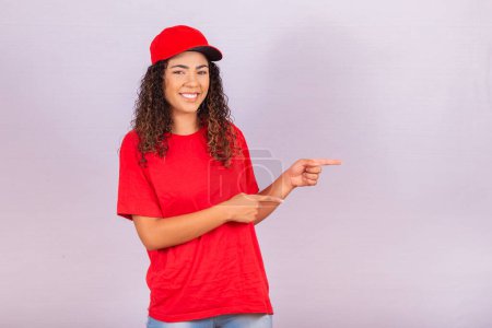 Téléchargez les photos : Livraison employée femme en chapeau rouge blanc t-shirt uniforme de travail messager en service pendant la quarantaine coronavirus covid-19 virus, pointant les doigts de côté sur l'espace de travail isolé sur fond blanc studio - en image libre de droit