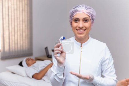 Téléchargez les photos : Gros plan sur esthéticienne tenant microneedling sourire à une caméra avec le patient en arrière-plan - en image libre de droit