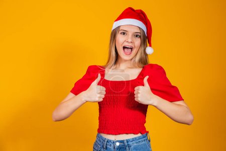 Téléchargez les photos : Portrait de femme joyeuse montrant pouces vers le haut portant le chapeau du Père Noël. - en image libre de droit