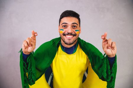 Téléchargez les photos : Fan brésilien avec un drapeau sur le dos acclamant avec les doigts croisés pour que le Brésil gagne - en image libre de droit