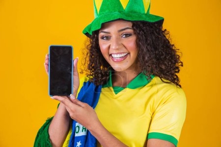 Téléchargez les photos : Ventilateur brésilien tenant un smartphone avec espace de copie. brésilien supporter féminin avec téléphone portable avec écran blanc pour l'écriture de texte. - en image libre de droit