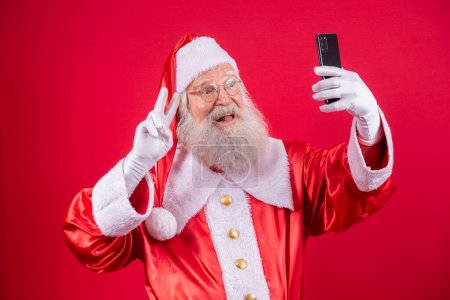 Photo for Stylish trendy grandfather aged mature Santa tradition winter costume headwear spectacles white beard take christmastime selfie picture front camera in red background. - Royalty Free Image
