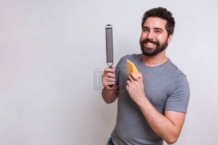 Photo for Young man holding a grater and a piece of cheese in his hand on gray background. man will grate the cheese - Royalty Free Image