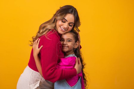 Photo of a beautiful young mommy holding her arms hugging her little daughter good mood adorable sincere heart feelings in the back jeans t-shirts isolated with yellow color background