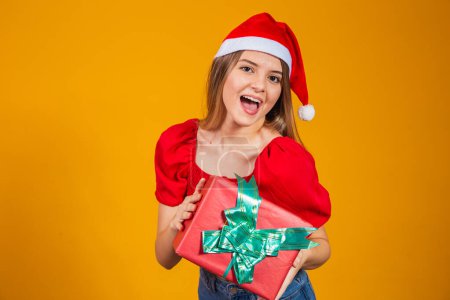 Téléchargez les photos : Femme excitée souriante en tenue rouge Père Noël tenant cadeau de Noël à son oreille isolée sur le fond jaune - en image libre de droit