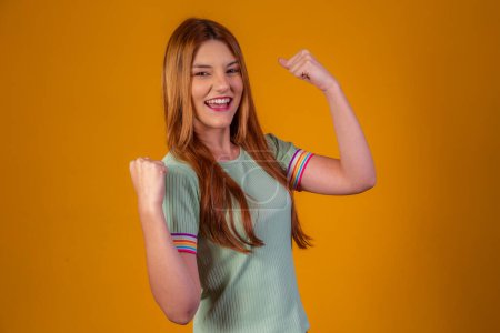 Happy and excited beautiful young woman expressing winning gesture. Successful victory and celebrating, triumphant, yellow background