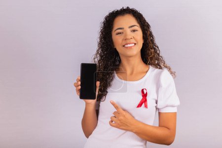 Foto de El concepto del Día Mundial del SIDA. Mujer joven sostiene teléfono inteligente con pantalla en blanco y cinta roja en la camiseta con el concepto de prevención y programación del VIH. - Imagen libre de derechos