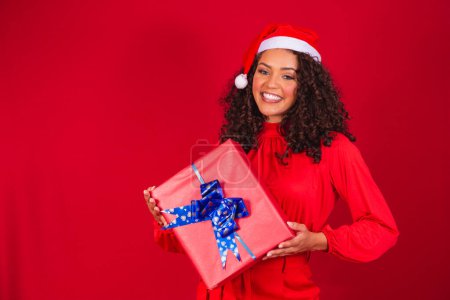 Téléchargez les photos : Portrait de jeune femme afro au chapeau de Père Noël tenant un cadeau sur fond rouge. concept de nuit de Noël - en image libre de droit