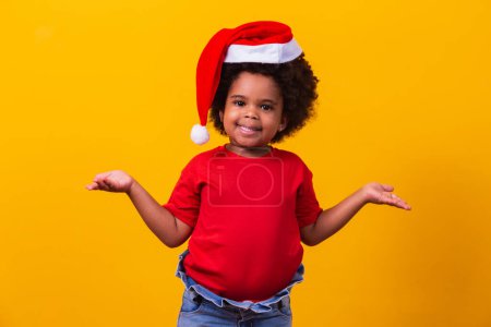 Téléchargez les photos : Petite Afro fille en T-shirt rouge et chapeau de Père Noël habillé pour Noël avec espace pour le texte pointant. - en image libre de droit