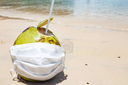 Téléchargez les photos : Photo d'une noix de coco sur la plage avec masque chirurgical. Concept de pandémie et de vacances - en image libre de droit