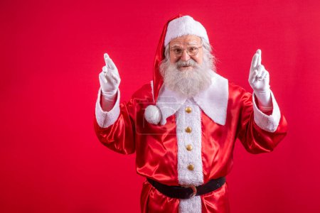 Téléchargez les photos : Portrait d'homme âgé rêveur en costume de Père Noël croisant les doigts faisant un voeu, magie de Noël, vacances d'hiver. Studio intérieur tourné isolé sur fond rouge - en image libre de droit