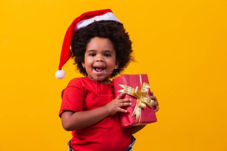 Téléchargez les photos : Enfant afro souriant au chapeau rouge du Père Noël tenant à la main un cadeau de Noël. Concept de Noël. - en image libre de droit
