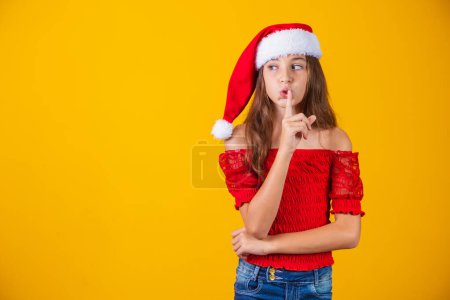 Téléchargez les photos : Petite fille habillée pour Noël faisant signe de silence sur fond jaune. - en image libre de droit