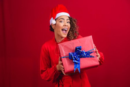 Foto de Retrato de una joven afro en sombrero de Santa Claus sosteniendo un regalo sobre fondo rojo. concepto de la noche de Navidad - Imagen libre de derechos